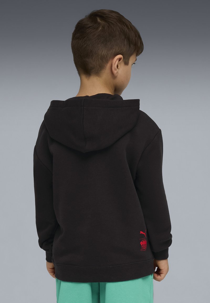 Garçon de dos portant un sweat à capuche noir avec un logo rouge en bas et un pantalon vert clair sur fond gris.