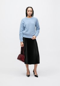 Hellblauer Strickpullover, schwarzer plissierter Midirock, rote Handtasche und schwarze High Heels. Fokus auf Textur und Farbkontrast im Outfit.