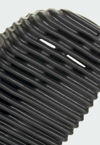 Svart gummimatta med en vågig textur och ribbat mönster, med spår för grepp och ventilation. Kanterna är rundade.