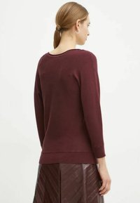 Maglione bordeaux con scollo tondo, maniche lunghe e polsini a coste. Il tessuto sembra morbido, abbinato a una gonna marrone elegante con motivo.