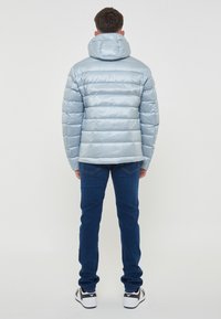 Giacca puffa azzurra con cappuccio, caratterizzata da un design trapuntato orizzontale, texture lucida e un retro leggermente più lungo. Indossata con jeans blu e sneaker.