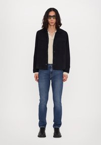 Veste noire en corduroy sur un pull beige, associée à un jean bleu et des chaussures noires. Des lunettes de soleil sombres complètent la tenue. Fond uni.