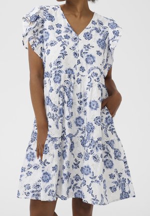 Vrouw draagt een witte jurk met blauw bloemmotief, korte gerimpelde mouwen, V-hals en hand in de zak.