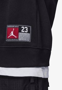 Sweatshirt preto com uma etiqueta apresentando o número "23" e um logótipo em silhueta. Sobreposto a uma camisa branca por baixo, com o tecido texturizado visível.