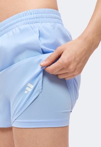 Main tirant un short de sport bleu clair pour révéler un short de compression intégré avec un logo blanc sur la cuisse.