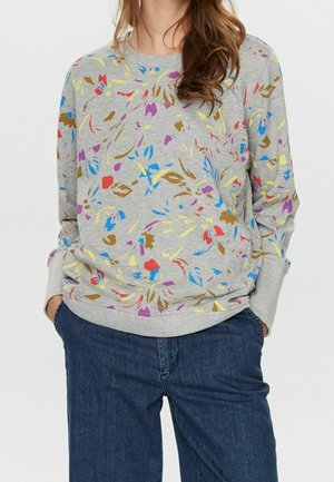 Femme portant un sweat-shirt gris avec des motifs floraux abstraits multicolores et un jean bleu, mains dans les poches, debout devant un fond uni.