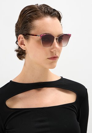 Donna con capelli corti castani che indossa occhiali da sole a forma di occhio di gatto color tartaruga e rosa e una maglia nera con un taglio sul petto.