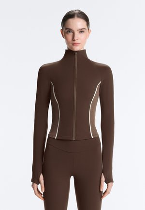 Donna che indossa una giacca marrone aderente con cerniera e cuciture laterali bianche, abbinata a leggings marroni, in piedi davanti a uno sfondo neutro.