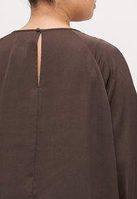 Brązowa bluzka z otworem w kształcie łezki z tyłu, wykonana z miękkiego materiału, z delikatnymi detalami przeszycia wzdłuż szwów.