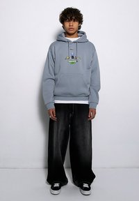 Szara bluza z kapturem z kieszenią z przodu oraz kolorowym graficznym wzorem. W połączeniu z czarnymi, szerokimi dżinsami i czarnymi trampkami.