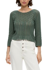 Grüner Strickpullover mit einer lockeren, atmungsaktiven Textur, weiter Boots-Ausschnitt und Dreiviertelärmeln, kombiniert mit hochtaillierten weißen Hosen mit Knöpfen.