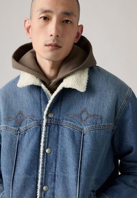 Denim jacket with a shearling collar, blue color, featuring brown stitching in a diamond pattern. Layered over a brown hoodie.