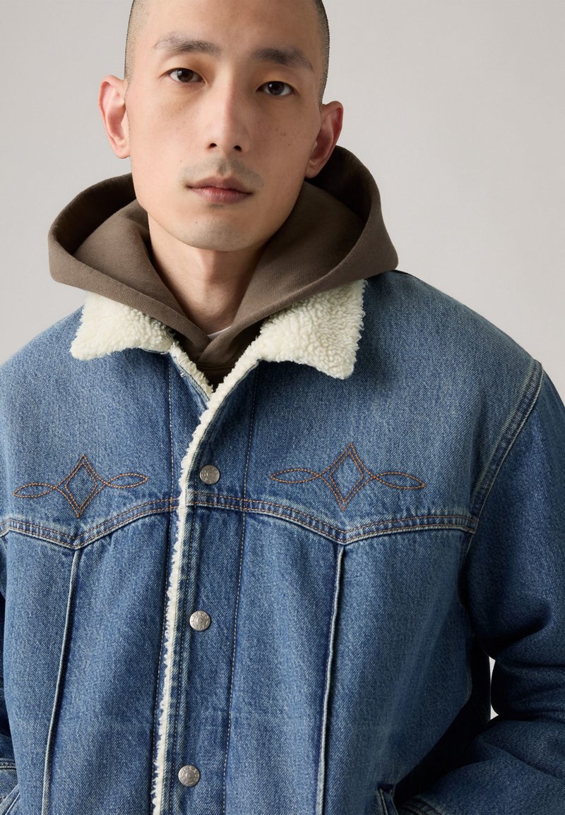 Dżinsowa kurtka z kołnierzem z owczej wełny, w kolorze niebieskim, z brązowymi szwami w wzór rombu. Nałożona na brązowy hoodie.