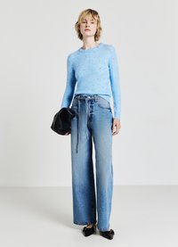 Pull en tricot bleu clair à pois, associé à un jean délavé à jambes larges. Le mannequin tient un sac à main noir et lisse. Des chaussures plates noires complètent le look.