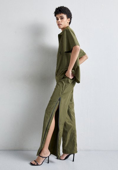 Olijfgroen outfit met een kortoliem shirt en rits brede broek. De stof lijkt lichtgewicht met een soepele textuur. Zwarte sandaaltjes met bandjes.