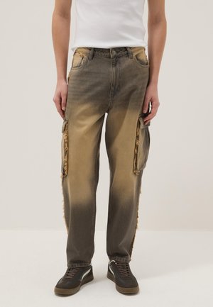 Persoon die vervaagde bruine cargobroek met versleten zijzakken en zwarte sneakers draagt, staand tegen een effen witte achtergrond.