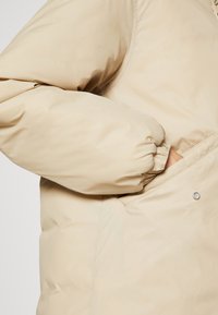 Chaqueta acolchada beige con una textura suave y lisa, que presenta un puño ajustado y un bolsillo lateral asegurado con un botón a presión.