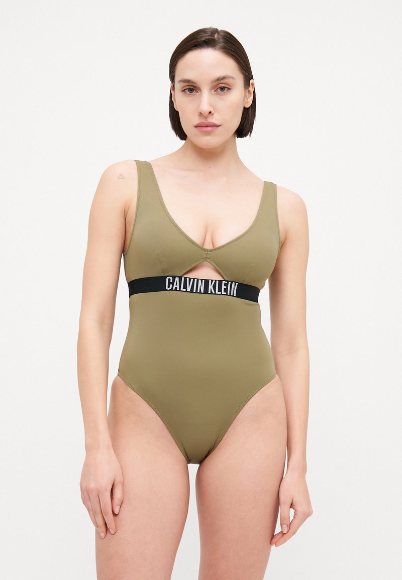 Olíva zöld egyrészes fürdőruha mély V-nyakkivágással és kivágott részlettel, fekete elasztikus szalaggal, rajta a "CALVIN KLEIN" logóval. Simító felület.