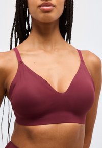Kvinne med flettet hår som bærer en sømløs dyp burgunder bralette med justerbare skulderstropper mot en ensfarget bakgrunn.