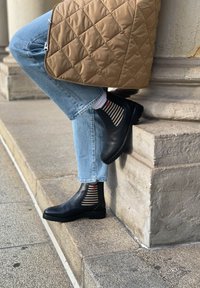 Bottes mi-hauteur en cuir noir avec des panneaux élastiques sur les côtés, associées à un jean en denim bleu clair et des chaussettes rayées avec un motif de cœur rouge.
