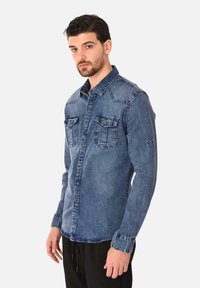 Camicia in denim azzurro medio con bottoni. Presenta due tasche sul petto, maniche lunghe e un colletto strutturato. Tessuto testurizzato con dettagli di cucitura.