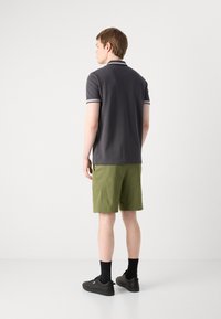 Pelēks polo krekls ar baltu apdari, apvienots ar olīvu zaļām šortiem. Modelis valkā melnus sporta apavus un zeķes, apskatīts no aizmugures.
