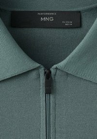 Prenda de punto verde con cremallera, cuello y etiqueta negra con la inscripción "PERFORMANCE MNG" en tamaño mediano para la UE, EE. UU. y México.