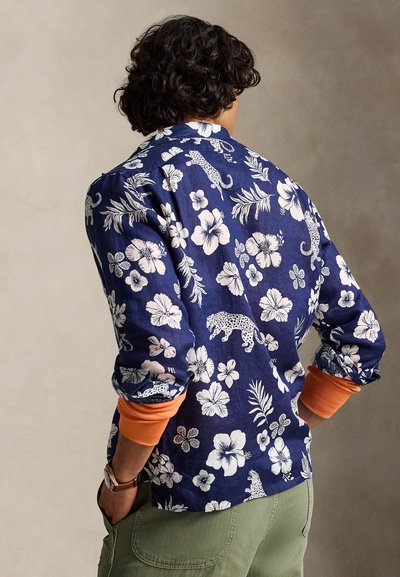 Personne aux cheveux bouclés et foncés portant une chemise bleue à motif floral et léopard sur un haut orange à manches longues, avec un pantalon vert et les mains dans les poches.