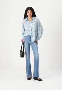 Camisa semitransparente de fundo azul claro com padrão e fecho de botões, combinada com jeans desbotados e em pé de flared. Uma mala preta e sapatos planos completam o look.