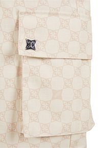 Mønstret stof i beige nuancer med et gentagende blomsterdesign. Omfatter en lomme med en dekorativ hardware-accent i sølvfarve.