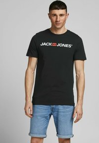 Schwarzes Baumwoll-T-Shirt mit weißem "JACK & JONES"-Logo, Rundhalsausschnitt, kurze Ärmel. Kombiniert mit hellblauen, aufgerollten Jeansshorts.