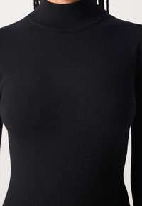 Gros plan d’une personne portant un pull à col roulé noir ajusté avec des cheveux tressés visibles au niveau du cou.