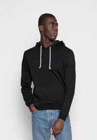 Sudadera negra con bolsillo frontal, capucha ajustable y puños de canalé. Llevada con vaqueros azul claro, que presentan una textura suave y un ajuste casual.