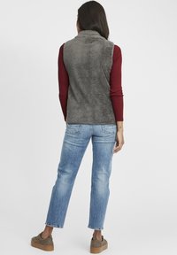 Grau Fleece-Weste mit hohem Kragen, kombiniert mit einem bordeauxfarbenen Langarmshirt. Helle Jeans und gemusterte Schuhe runden den Look ab.