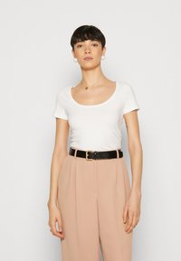 Camiseta blanca de manga corta con cuello redondo, combinada con unos pantalones de cintura alta color beige y un cinturón negro con hebilla dorada.