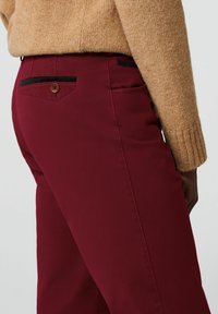 Bordeauxrode broek met een gladde textuur, voorzien van een achterzak met een bruine knoop en zwarte sierstiksels. Draagt een beige trui erbij.