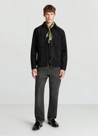 Veste en daim noir avec deux poches avant, portée sur une chemise noire, associée à un jean gris et des mocassins noirs. La tenue est agrémentée d'un foulard en soie vert.