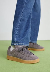 Grijze suède sneakers met rubberen gomzolen, voorzien van drie laterale strepen, een ronde neus en platte veters. Gecombineerd met blauwe spijkerbroeken.