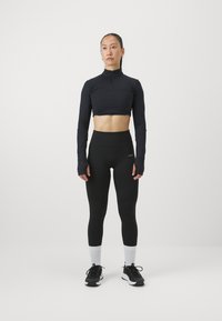 Haut noir à manches longues et cropped avec un col montant, associé à un legging noir taille haute. Le modèle porte des baskets noires avec des accents blancs et des chaussettes.