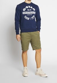 Mörkblå bomullströja med vitt grafiskt logo, i kombination med olivgröna shorts. Sammanfattningsdetaljer inkluderar rund hals och ribbade muddar.