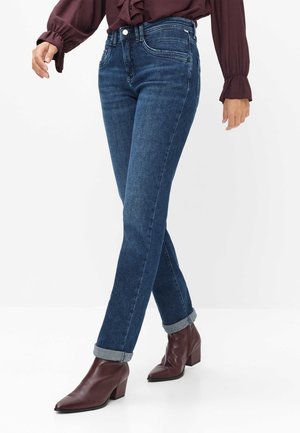 Person iført mørkeblå opsmøgede jeans, burgunderfarvede hælede ankelstøvler og en mørkelilla langærmet bluse, der går.
