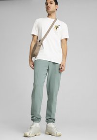 Camiseta de algodón blanca con un estampado gráfico, pantalones de chándal verde claro con puños de jogger y zapatillas beige. Se lleva un bolso cruzado beige.