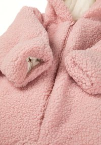 Manteau en polaire rose avec une surface douce et texturée, doté d'une fermeture éclair et de poches surdimensionnées et arrondies. Le matériau a l'air pelucheux et confortable.