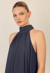 Bahdittumadiinen halter jätti sinisessä kankaassa, jossa on korkea kaula, laskostettu muotoilu ja avoimet olkapäät. Minimalistinen tyyli, jossa on sileä texture.