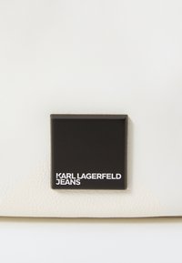 Krém színű bőr táska, textúrált felülettel, fekete négyzet alapú logó táblával, amelyen fehér betűkkel az "KARL LAGERFELD JEANS" felirat található.