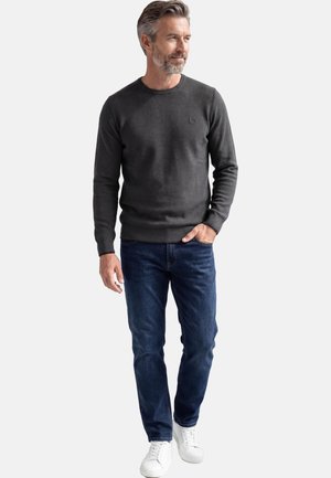 Mann mit grauen Haaren, der einen dunkelgrauen Pullover, blaue Jeans und weiße Turnschuhe trägt, steht mit einer Hand in der Tasche und blickt nach rechts.