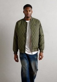 Veste bomber matelassée vert olive avec devant zippé, ourlet et poignets côtelés, portée sur une chemise blanche, associée à un jean bleu.