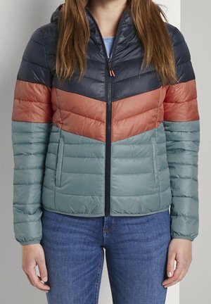 Kvinde iført en lystæppet jakke med lynlås med navy, rust og gråblå farveblokke, kombineret med blå jeans, stående mod en neutral baggrund.