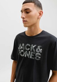 Sort bomuld T-shirt med rund halsudskæring, med store, teksturerede bogstaver "JACK & JONES" i et bladmønstret design på forsiden.