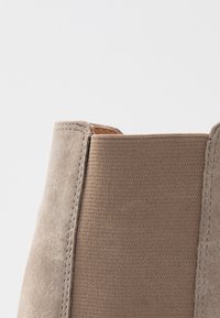 Botín de ante y tela con un color beige, acabado texturizado y costuras visibles a lo largo de las uniones. No presenta detalles metálicos.